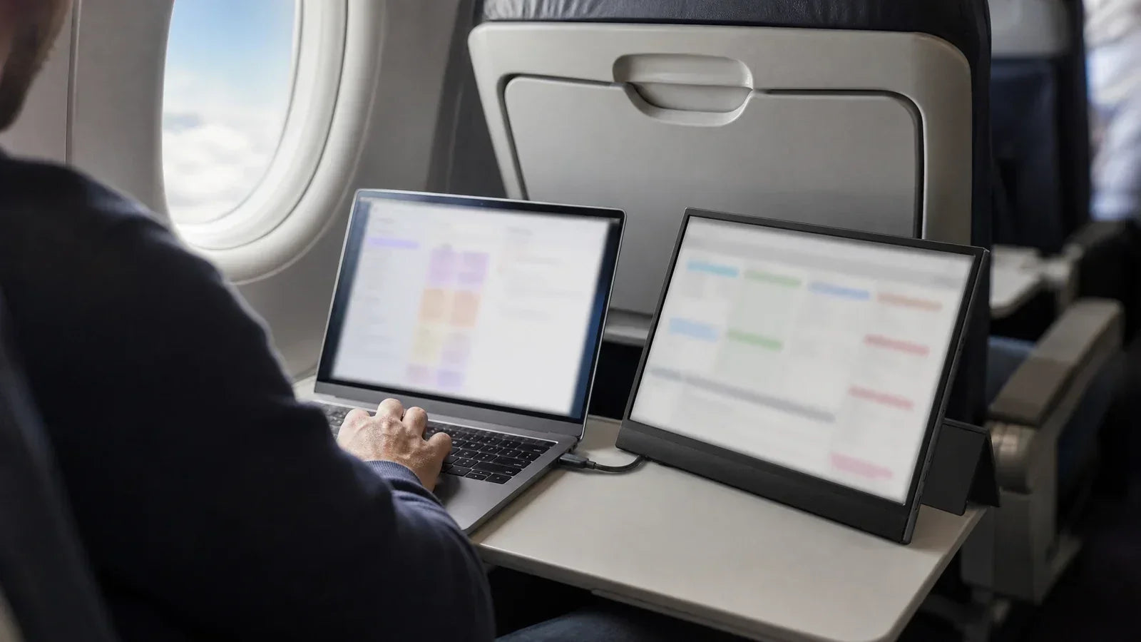 Laptop and portable travel monitor set up side by side on an aeroplane tray table beside the cabin window